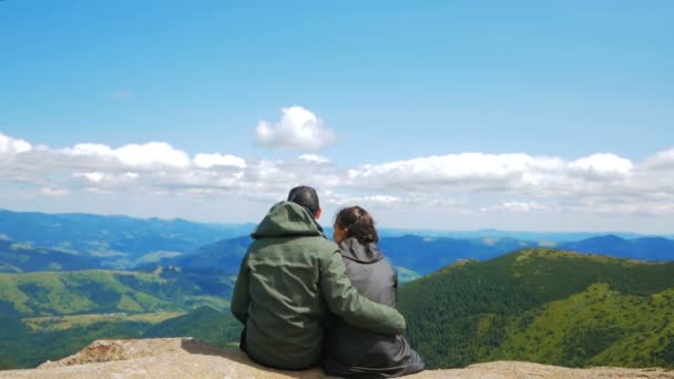Birbirine aşık genç turistler bir uçurumun tepesinde oturup ihtişamlı dağların arka planına tutkuyla öpüşürler. Genç çekici kadın gezgin ve genç çekici — Stok video