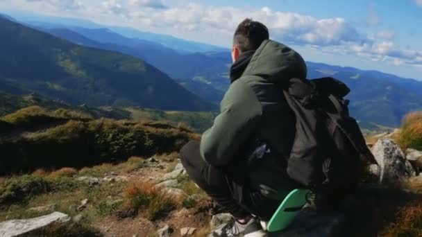 Young guy traveler enjoys the view of the mountain range while sitting on the top of the cliff. — Stock Video