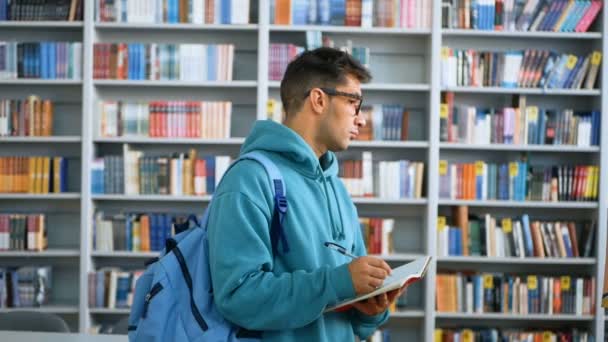 Молодая студентка тысячелетия пишет в блокноте, стоя в публичной библиотеке на фоне книжных полок. — стоковое видео