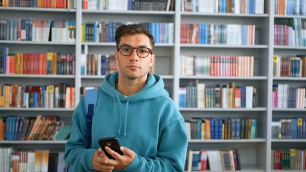 Gözlüklü genç bir milenyum öğrencisi, kütüphanenin arka planında kitaplıkların karşısında dururken akıllı telefonuyla sosyal ağda geziniyor.. — Stok video
