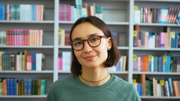 Retrato de cerca de una joven estudiante en el contexto de las estanterías de la biblioteca universitaria. Joven mujer milenaria mirando a la cámara y sonriendo mientras está parada en una biblioteca pública — Vídeos de Stock