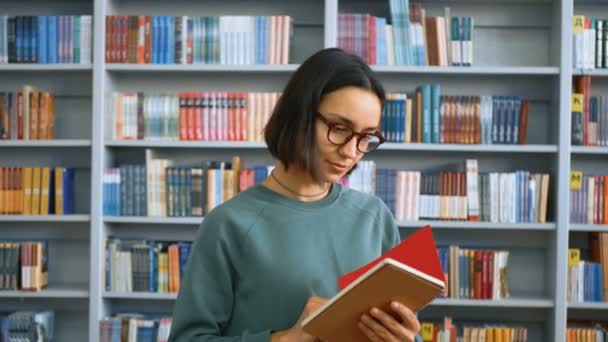 Uma jovem estudante milenar escreve uma tarefa em seu caderno enquanto está em uma biblioteca pública no contexto de estantes de livros. Retrato de close-up de uma jovem estudante atraente — Vídeo de Stock