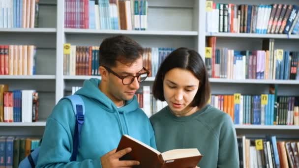 Eine junge Jahrtausendstudentin mit Brille und eine junge attraktive Studentin lesen ein Buch und bereiten sich auf eine Prüfung vor, während sie in einer öffentlichen Bibliothek vor dem Hintergrund von Bücherregalen stehen.. — Stockvideo