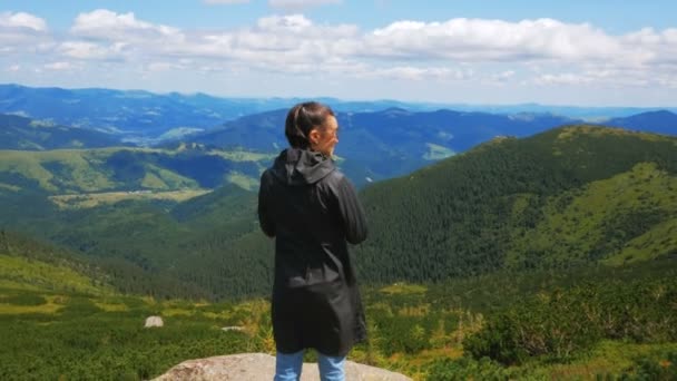 Uma jovem viajante fica no topo do penhasco e olha para a paisagem da montanha. Uma jovem mulher atraente viajante fica no topo da montanha e olha para a distância na — Vídeo de Stock