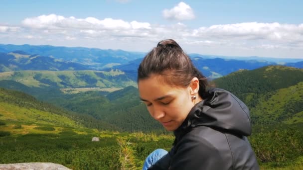 若いハイカーの女性と笑みを浮かべて、カメラの崖の上の山の風景の背景に座って見て。若い女性旅行者が山の頂上に到達し、美しいを楽しんでいます — ストック動画