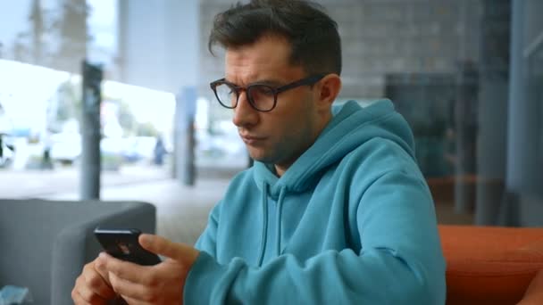 Νέος ελκυστικός επιχειρηματίας διαβάζει τις ειδήσεις σε ένα κοινωνικό δίκτυο στο smartphone του, ενώ κάθεται σε μια καρέκλα. — Αρχείο Βίντεο