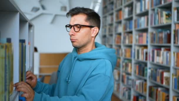 De cerca retrato de un joven estudiante atractivo que está buscando el libro correcto en la estantería de la biblioteca universitaria.Un joven estudiante milenario con gafas selecciona un libro interesante sobre el — Vídeos de Stock