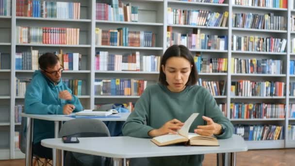 Junge attraktive Universitätsstudenten bereiten sich auf den Test vor, sie sitzen an den Tischen in der öffentlichen Bibliothek und schreiben die nötigen Informationen in Notizhefte.. — Stockvideo
