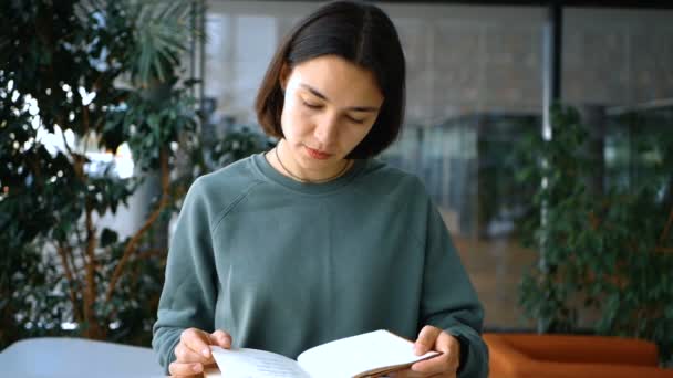 Νεαρή ελκυστική φοιτήτρια διαβάζει ένα βιβλίο και κρατά σημειώσεις σε ένα σημειωματάριο, ενώ ετοιμάζεται για εξετάσεις, ενώ κάθεται σε ένα τραπέζι σε μια δημόσια βιβλιοθήκη. Κοντινό πλάνο πορτρέτο μιας νεαρής χαρούμενης γυναίκας — Αρχείο Βίντεο