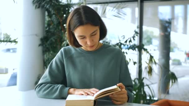 Genç çekici kız öğrenci bir kitap okuyor ve halk kütüphanesinde bir masada otururken ne okuduğunu düşünüyor. Genç, neşeli bir kız öğrencinin yakın plan portresi. — Stok video