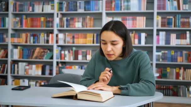 Νεαρή ελκυστική φοιτήτρια διαβάζει ένα βιβλίο και κρατά σημειώσεις σε ένα σημειωματάριο, προετοιμάζεται για μια εξέταση, ενώ κάθεται σε ένα τραπέζι σε μια δημόσια βιβλιοθήκη με φόντο τα ράφια των βιβλιοθηκών. Κοντινό πλάνο πορτρέτο του — Αρχείο Βίντεο