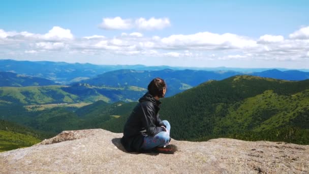 En ung kvinna resenär har nått toppen av berget och har en vacker utsikt över skogen och bergen. Ung vandrare kvinna ler och tittar på kameran sitter på toppen av en klippa på — Stockvideo