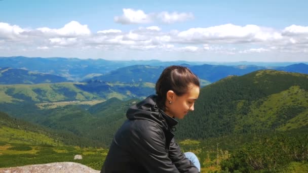 Uma jovem viajante chegou ao topo da montanha e desfruta de uma bela vista da floresta e das montanhas. Jovem caminhante mulher sorrindo e olhando para a câmera sentada no topo de um penhasco no — Vídeo de Stock