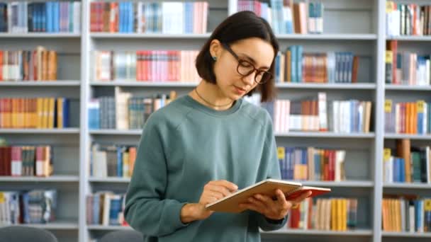 Nahaufnahme einer jungen attraktiven Lehrerin, die eine Lektion erteilt und die Aufgabe in einem Notizbuch vor dem Hintergrund der Bücherregale in der Universitätsbibliothek liest. — Stockvideo