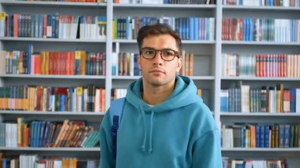 Retrato de cerca de un joven estudiante atractivo en el fondo de las estanterías de la biblioteca de la universidad. Joven estudiante milenario mirando a la cámara y sonriendo mientras está de pie en un público — Vídeos de Stock