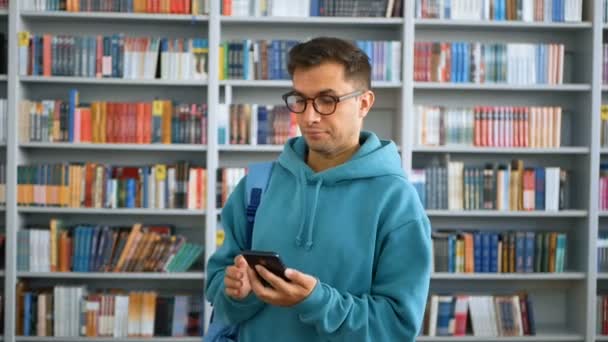 若い魅力的な学生の男のクローズアップ肖像,彼は大学図書館の本棚の背景に彼のスマートフォン上の友人とテキストメッセージを送っています.. — ストック動画