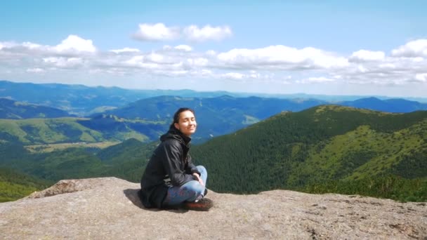 若い女性旅行者が山の頂上に到達し、森と山の美しい景色を楽しんでいます。若いハイカーの女性笑顔とカメラを見て崖の上に座っている — ストック動画