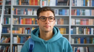 Genç öğrenci milenyumu, gözlükleriyle kameraya bakıyor ve kütüphanede arka planda kitap raflarına yaslanırken gülümsüyor. Eğitim kavramı.