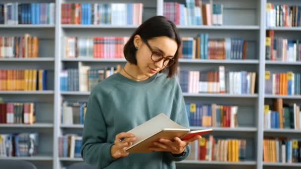若い女性の千年生が書棚を背景に公共図書館に立ってノートに課題を書きます。教育理念. — ストック動画