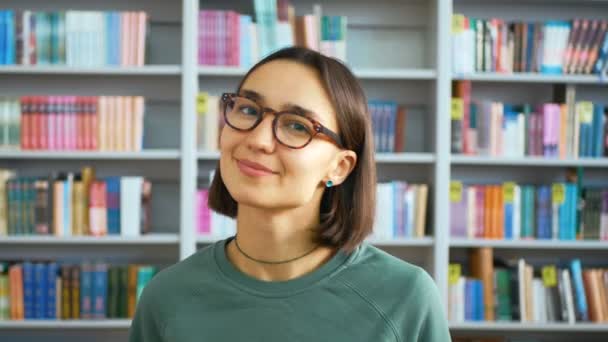 Νεαρή γυναίκα της χιλιετίας κοιτάζει την κάμερα και χαμογελά ενώ στέκεται σε μια δημόσια βιβλιοθήκη στο παρασκήνιο των ραφιών βιβλίων.Κοντινό πλάνο πορτρέτο μιας νεαρής φοιτήτριας στο φόντο του — Αρχείο Βίντεο