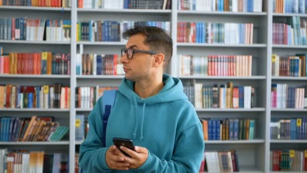 Um jovem estudante millennial com óculos vira através da rede social alimentar em seu smartphone, enquanto está em uma biblioteca pública contra o fundo de estantes. Retrato de close-up de um jovem — Vídeo de Stock