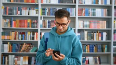 Genç ve çekici bir öğrencinin yakın plan portresi, üniversite kütüphanesindeki kitaplıkların arka planında akıllı telefonuyla arkadaşlarıyla mesajlaşıyor. Eğitim kavramı.