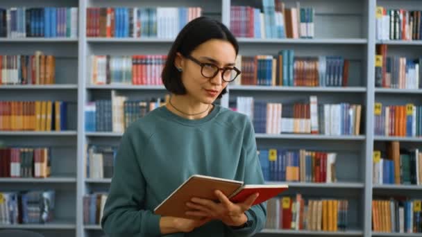 Ritratto ravvicinato di una giovane insegnante attraente che insegna una lezione, leggendo l'incarico in un quaderno sullo sfondo delle librerie della biblioteca universitaria. Concetto educativo. — Video Stock