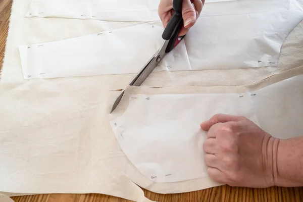 Designer's hand cuts calico cloth by scissors according with paper layouts of dress on wooden table at home — Stock Image Designer Hand Cuts Calico Cloth Scissors According Paper Layouts Dress Stock Image