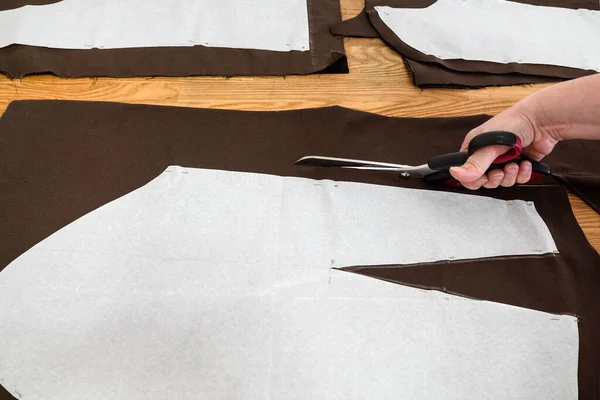 Woman's hand cuts brown fabric by scissors according with layouts of sewing patterns of dress on wooden table at home — Stock Image Woman Hand Cuts Brown Fabric Scissors According Layouts Sewing Patterns Stock Picture