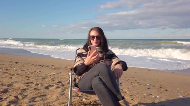 Брунетт говорить по мобільному телефону. жінка з темним волоссям сидить на піщаному пляжі — стокове відео