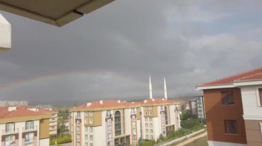 Gökyüzündeki kara bulutlar. Uzakta yüksek binalar var. Gökyüzünde büyük parlak gökkuşağı