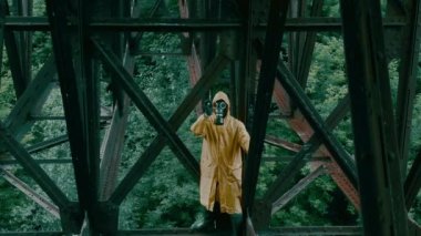 Koruyucu kıyafetli, demir köprünün altında gaz maskeli bir adam elini sallıyor.