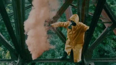 Koruyucu giysili ve gaz maskeli adam köprünün altında duruyor ve elinde sis bombası sallıyor..