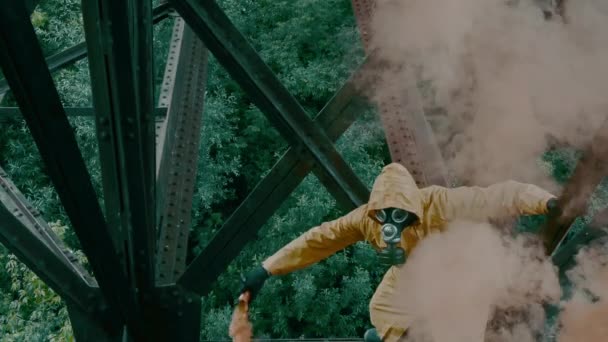 Homem de terno protetor e máscara de gás está sob ponte, brandindo bomba de fumaça. — Vídeo de Stock