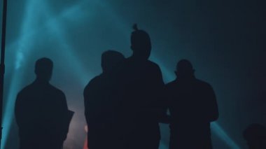 Mavi ışınlarla aydınlatılmış karanlık bir hangarda arkadan siyah erkek siluetleri..