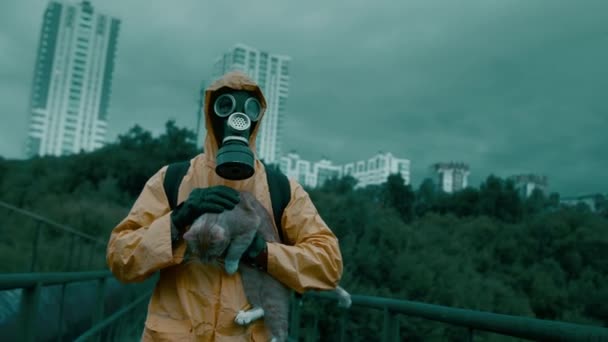 Man i gasmask, skyddsdräkt bär en katt i famnen mot. — Stockvideo