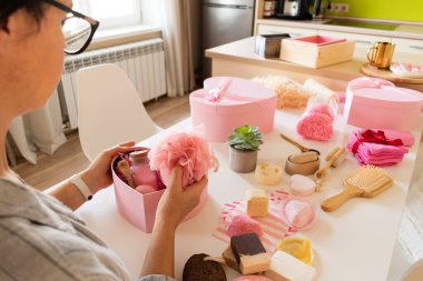 Doğal vücut bakımı, banyo malzemeleri, el yapımı sabun, banyo bombası, ahşap fırça, sünger taşı, sünger taşı, havlu ve dekoratif ayıyla seti hazırlayan bir kadın..