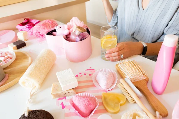 Woman preparing gift box set with natural body care and bath supplies of handmade soap, bath bombs, wooden brush, sponge, pumice stone, towels and decorative bear while having water with marshmallow.