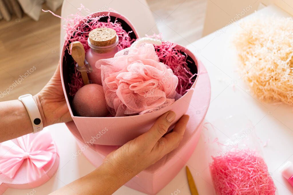 Woman preparing gift box set with natural body care and bath supplies of handmade soap, bath bombs, wooden brush, sponge, pumice stone, towels and decorative bear while having water with marshmallow.
