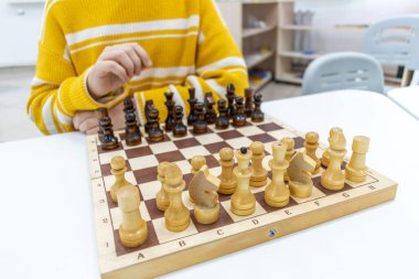 Çizgili sarı kazaklı kız tahta satranç tahtasında satranç oynuyor ve elinde satranç figürleri var. Boş zaman ve hobi anlayışı. Okul satranç kulübüne giden bir çocuk. Eğitim ve Geliştirme.