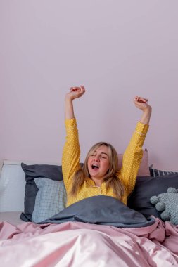 Genç bir kadın rahat bir yatakta otururken esniyor. Evim gibi. Refah kavramı.