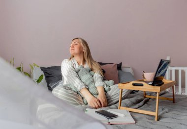 Genç iş kadını, işten sonra internette bilgisayar kullanarak yatakta otururken meditasyon yapıyor. Kız dizüstü bilgisayarın yanında sakin müzik dinliyor. Ev konfor bölgesi ve sağlık konsepti.