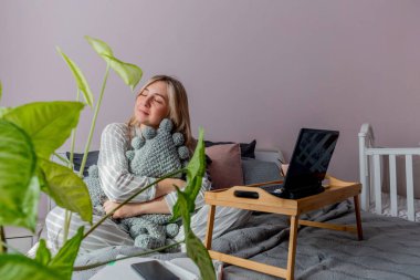 Genç iş kadını, işten sonra internette bilgisayar kullanarak yatakta otururken meditasyon yapıyor. Kız dizüstü bilgisayarın yanında sakin müzik dinliyor. Ev konfor bölgesi ve sağlık konsepti.