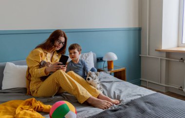 Genç anne ve oğlu arkadaşlarıyla video bağlantısı ile iletişim kuruyor. Cep telefonuyla konuşma. Psikolojik sağlık konsepti. Sabah yatakta.