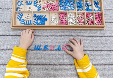 Top view of kids hands building words by using colored Montessori movable alphabet from the wooden tray on gray lined rug. Concept of learning practicing hand coordination and developing motor skills. clipart