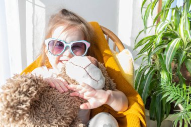 Defoktus. Küçük kız güneşin altında sallanan bir sandalyede oturur, kapalı alandaki bitkilerin arasında. Parlak gölge. Samimi insanlar konsepti. Evde dinlen.