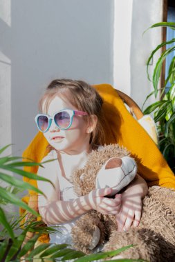 Defoktus. Küçük kız güneşin altında sallanan bir sandalyede oturur, kapalı alandaki bitkilerin arasında. Parlak gölge. Samimi insanlar konsepti. Evde dinlen.