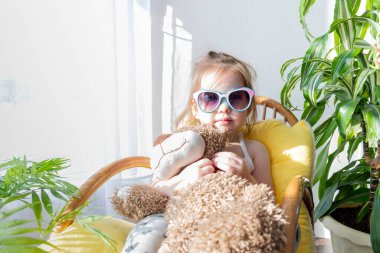 Defoktus. Küçük kız güneşin altında sallanan bir sandalyede oturur, kapalı alandaki bitkilerin arasında. Parlak gölge. Samimi insanlar konsepti. Evde dinlen.