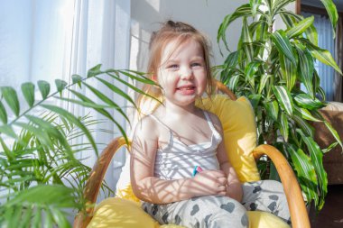 Defoktus. Küçük kız güneşin altında sallanan bir sandalyede oturur, kapalı alandaki bitkilerin arasında. Parlak gölge. Samimi insanlar konsepti. Evde dinlen.