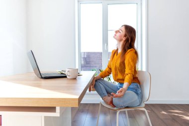 Genç iş kadını işten sonra internetten bilgisayar kullanarak meditasyon yapıyor. Dişi kız dizüstü bilgisayarın yanında sakin müzik dinler. Ev konforu ve ruh sağlığı konsepti.